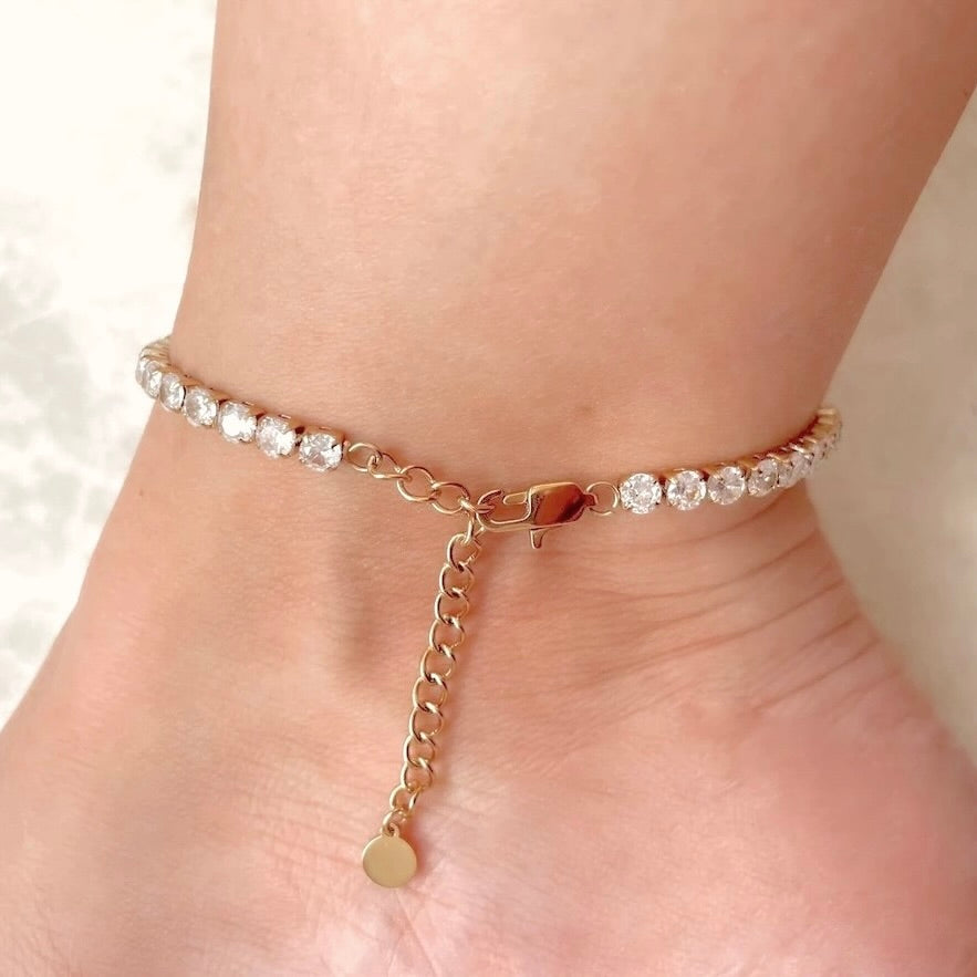 Gold tennis chain anklet with  a long lobster clasp, extender chain and tiny round stainless stamped tag at the end on a person's ankle against a neutral background, 