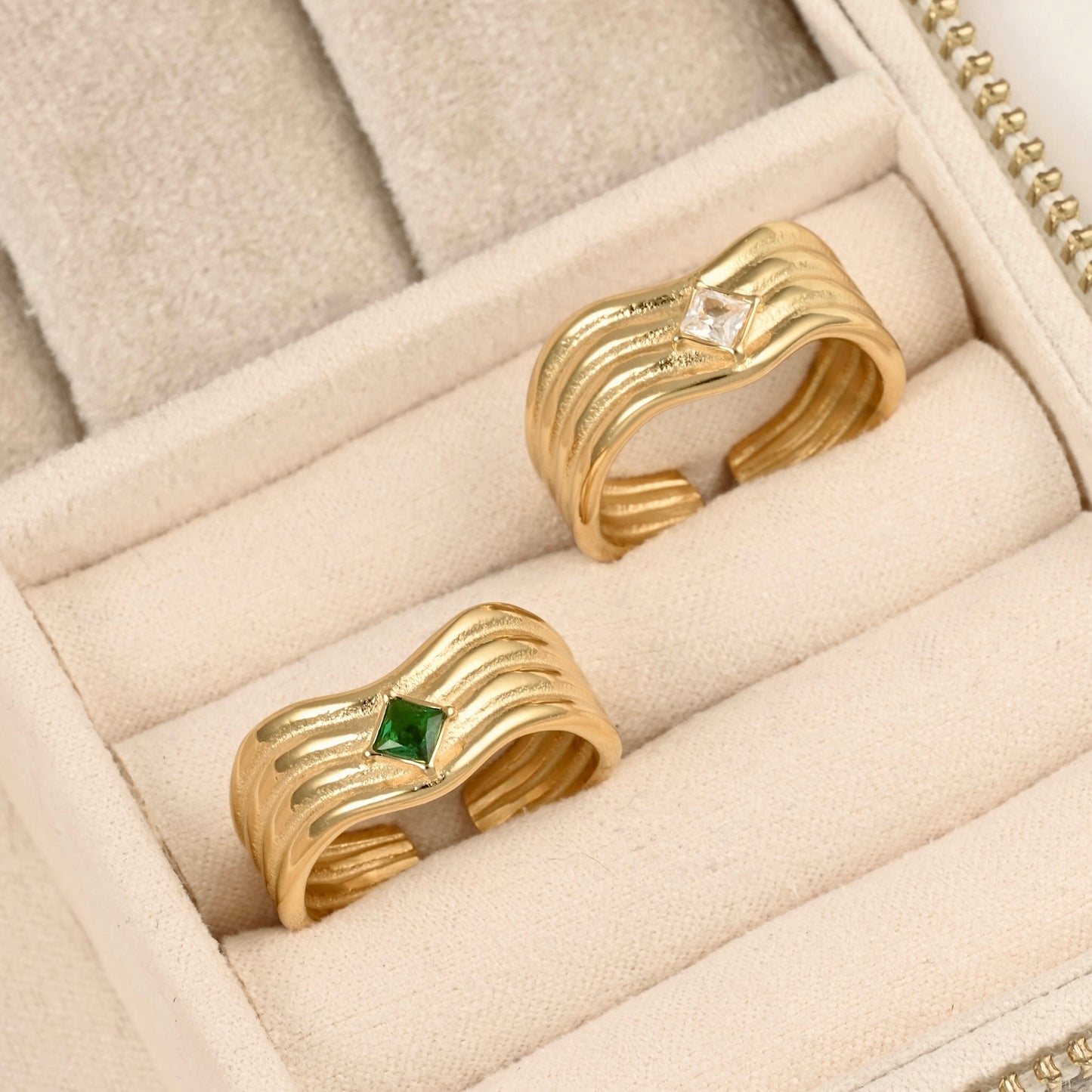 Two gold wave-shaped rings displayed in a beige jewelry box. One ring has a small square green gemstone, and the other has a small clear gemstone.