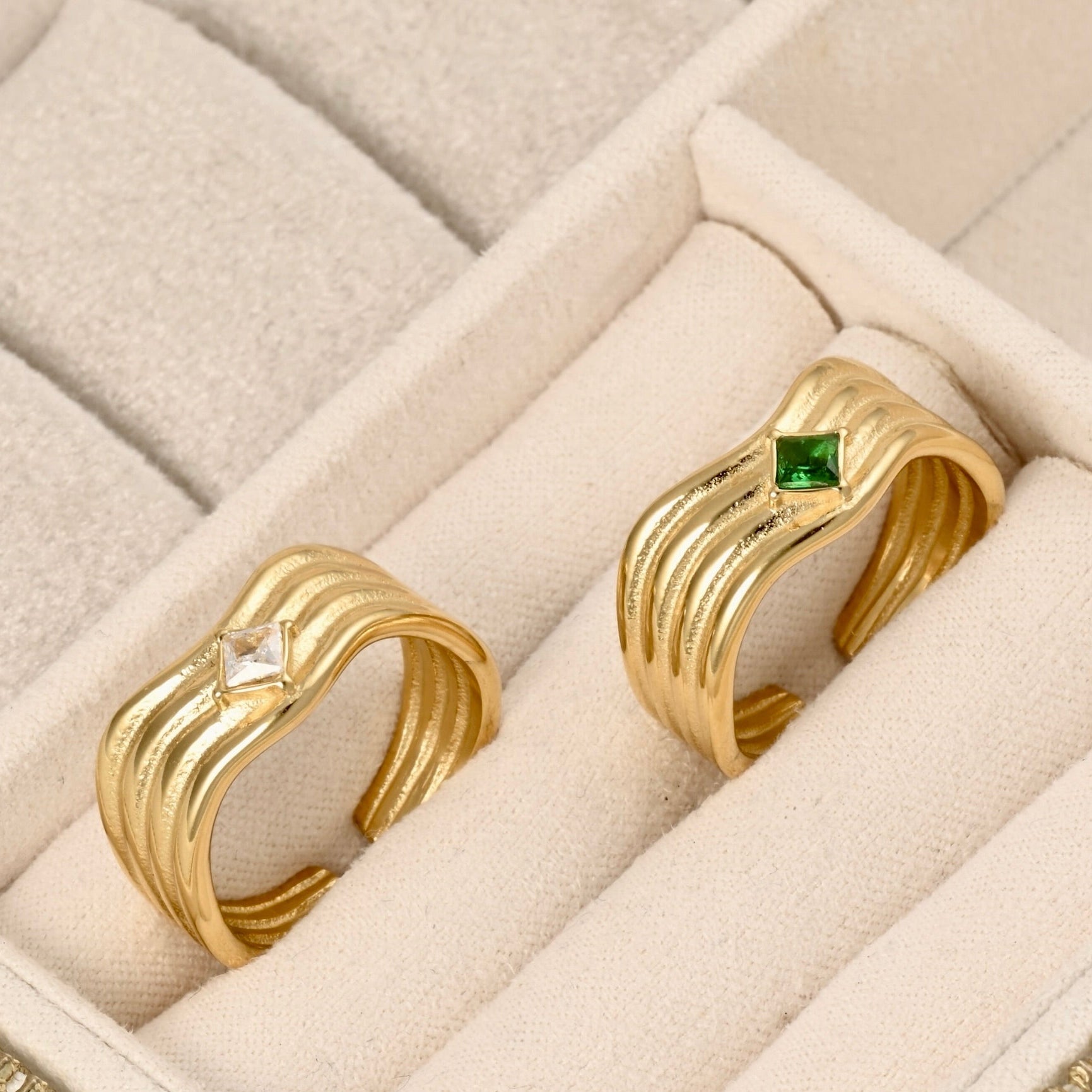 Two gold-toned wave-style rings displayed in a cream jewelry box. One ring has a small green square gemstone, and the other has a small clear diamond-shaped gemstone.