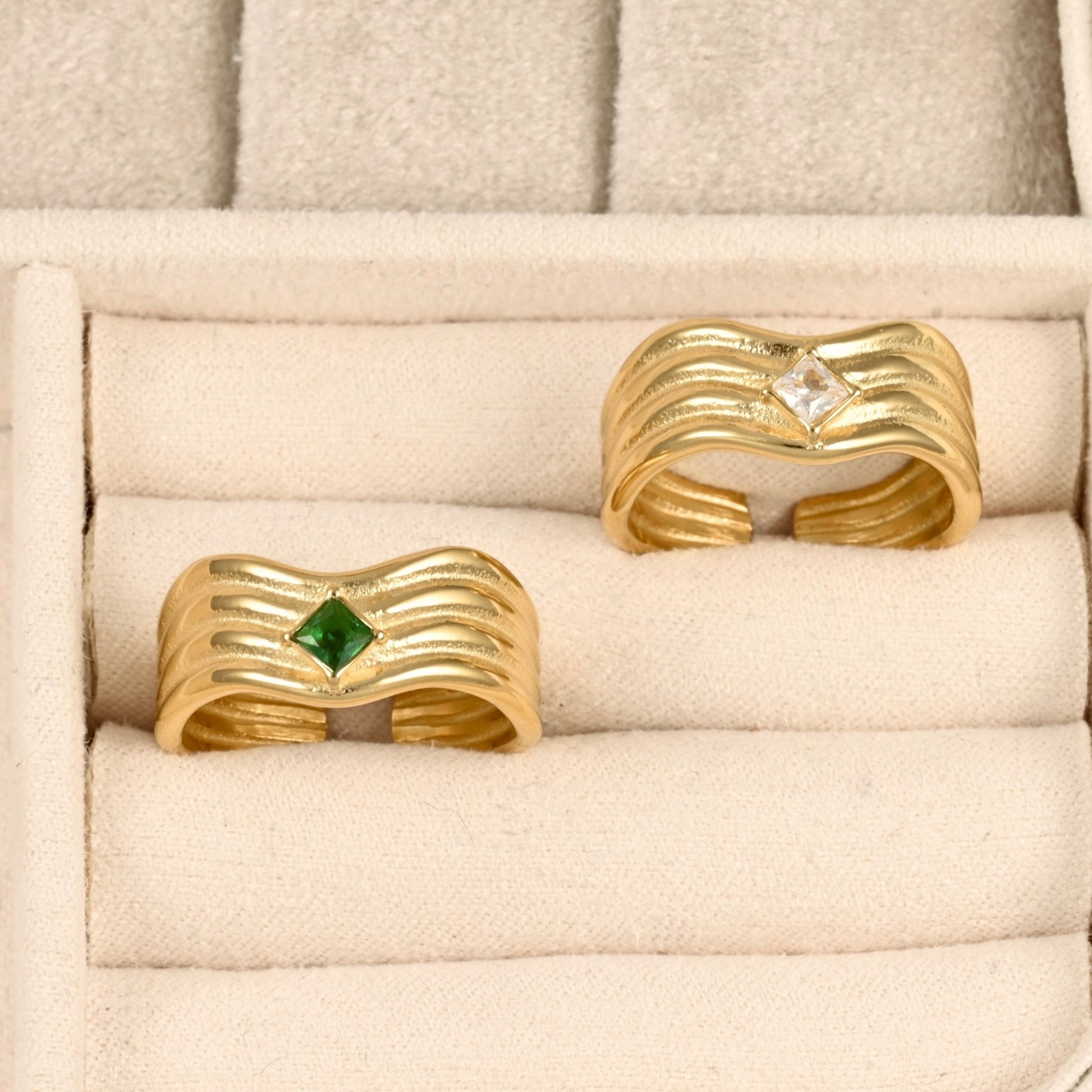 Two gold-colored wavy open-bend rings displayed in a cream-colored velvet jewelry box. One ring features a small green diamond-cut gemstone, and the other features a clear diamond-cut gemstone.