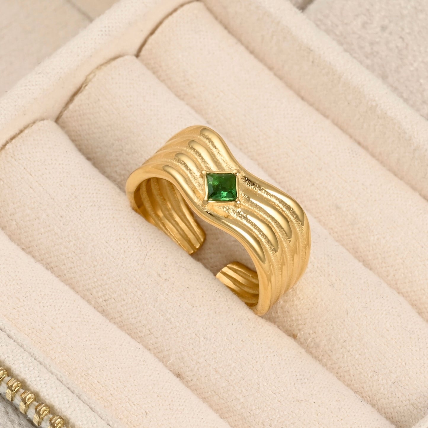 A gold ring with a wavy, ribbed band and a small green gemstone in the center, displayed inside a cream-colored jewelry box.