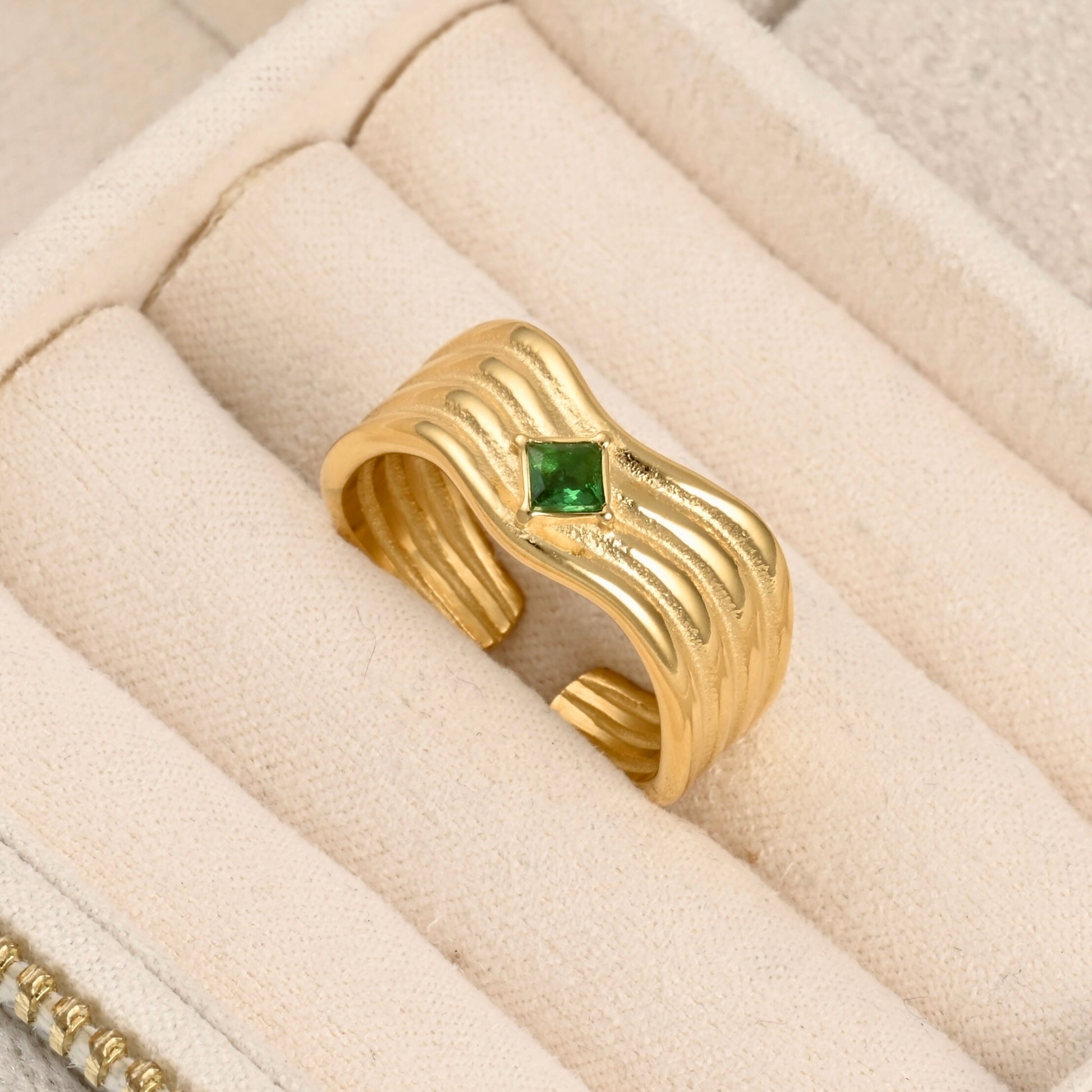 A gold ring with a wavy, ribbed band and a small green gemstone in the center, displayed inside a cream-colored jewelry box.