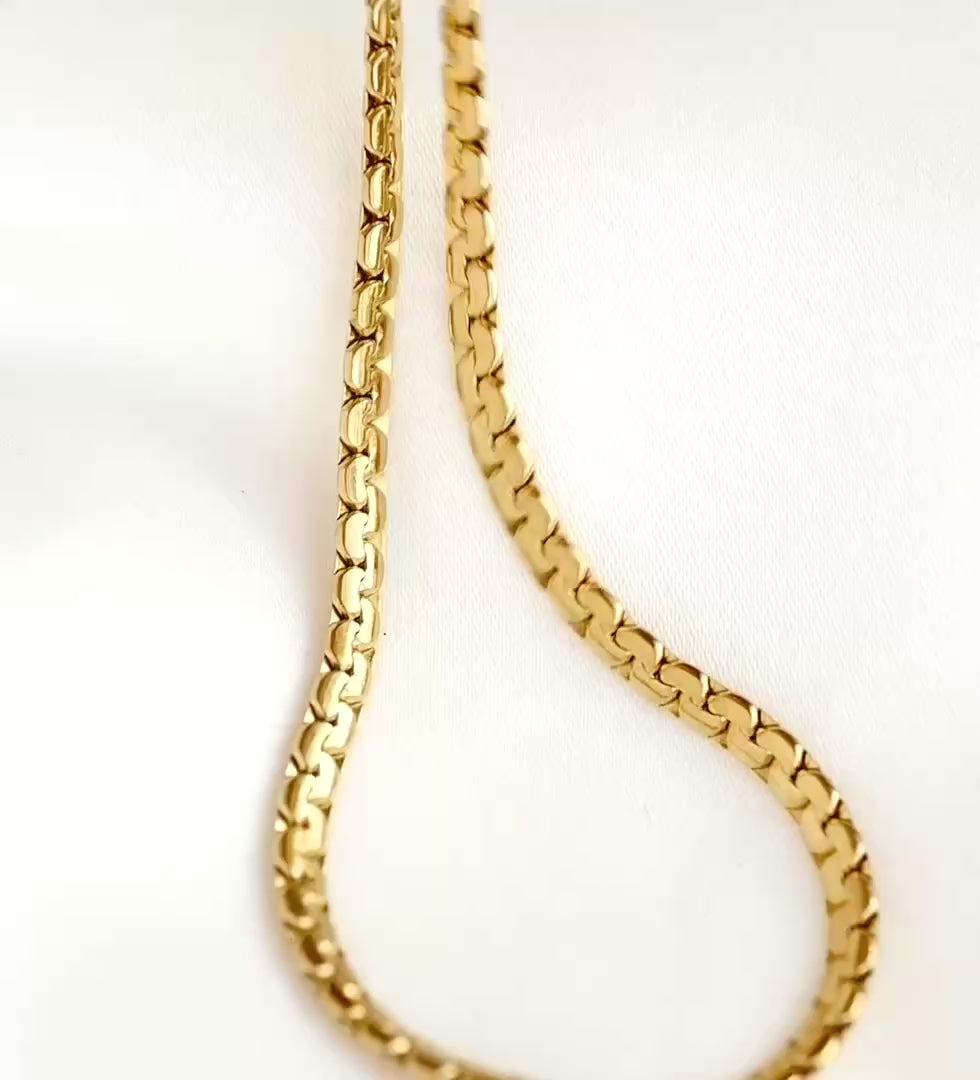 Gold snake bone chain on white satin fabric, the links reflecting light as the chain moves.