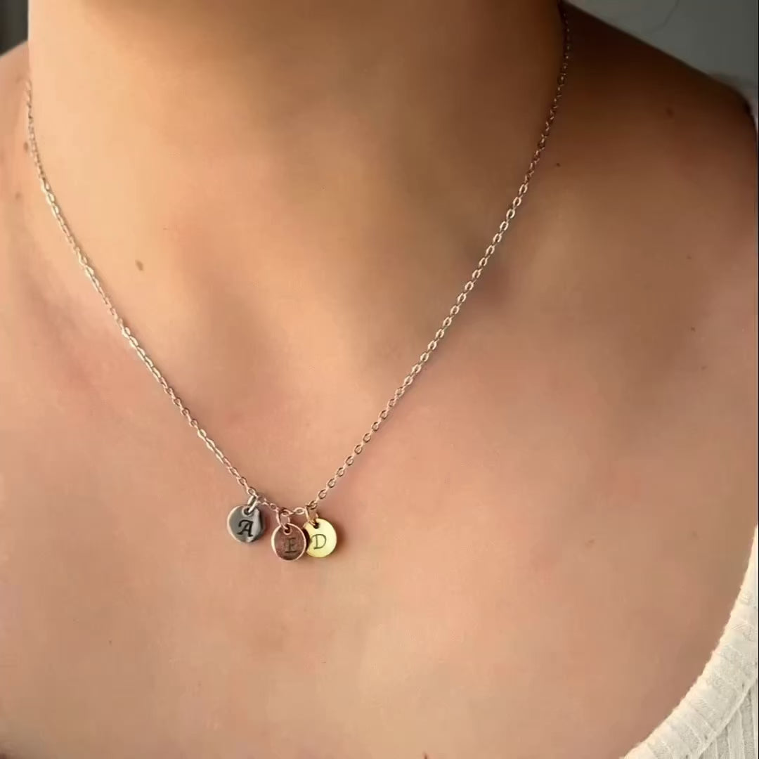 Person with white t-shirt wearing a silver cable chain necklace with gold, rose gold and silver initial discs which catch light as the person moves.