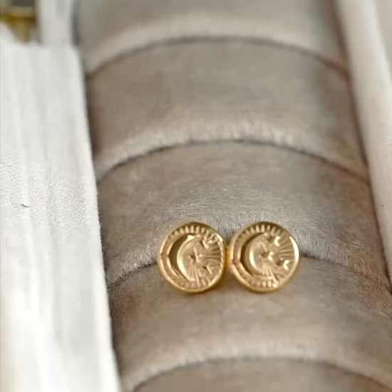 A pair of 18mm round coin stud earrings with an embossed crescent moon and 3 stars resembling a smiley face, the studs reflect light as the camera changes angle.
