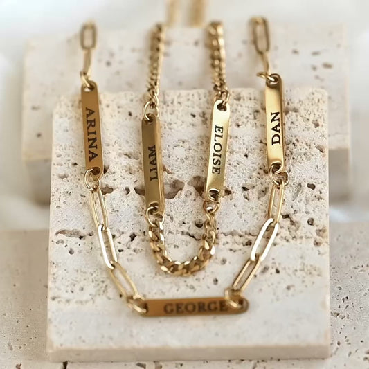 Two personalized gold nameplates on paperclip and curb chains, engraved with the names ARINA, WIL, ELOISE, DAN, and GEORGE, displayed on a textured beige stone block and name bars reflecting light as the reflector moves.