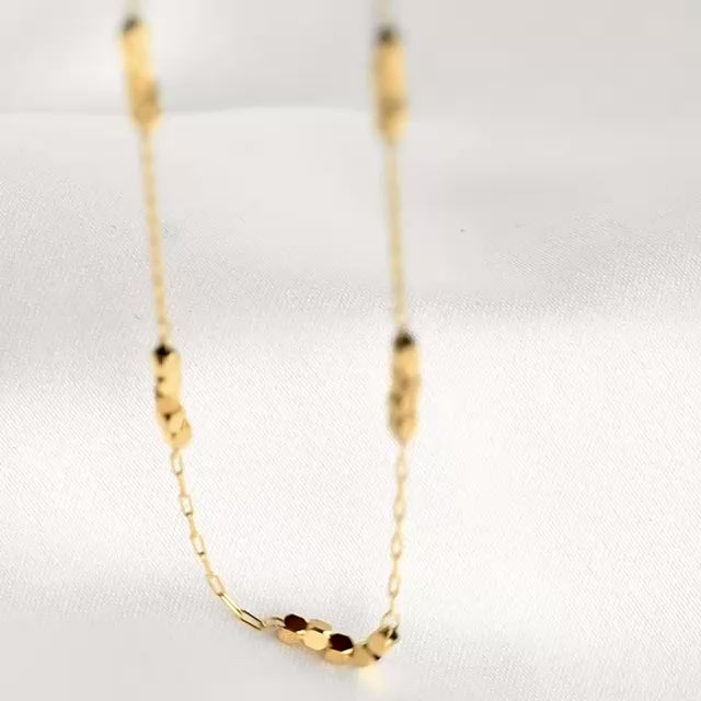 A delicate gold chain with cube beads against a white background, with gold beads reflecting light as chain moves 