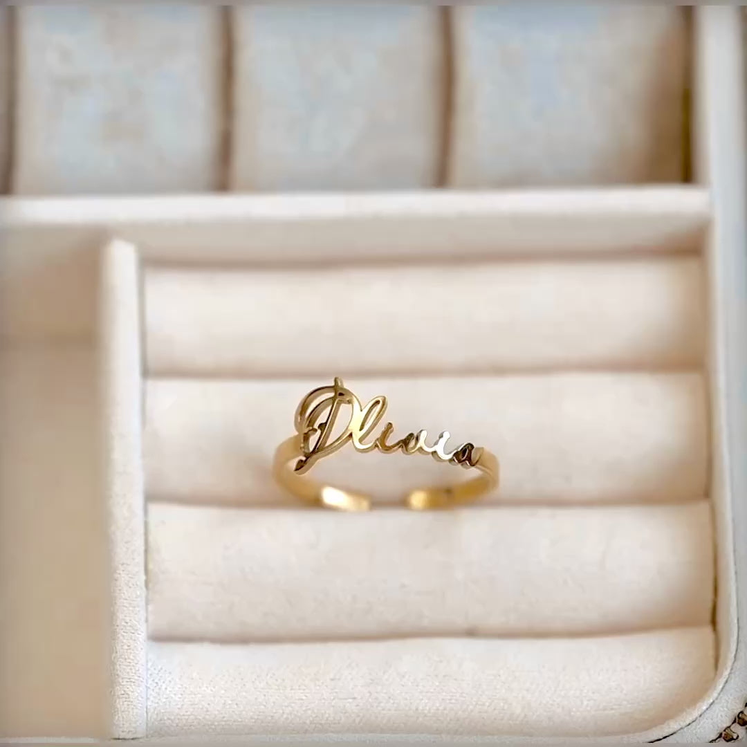 A gold name ring with 'Olivia' in a jewelry case, the letters reflecting light as the reflector moves.