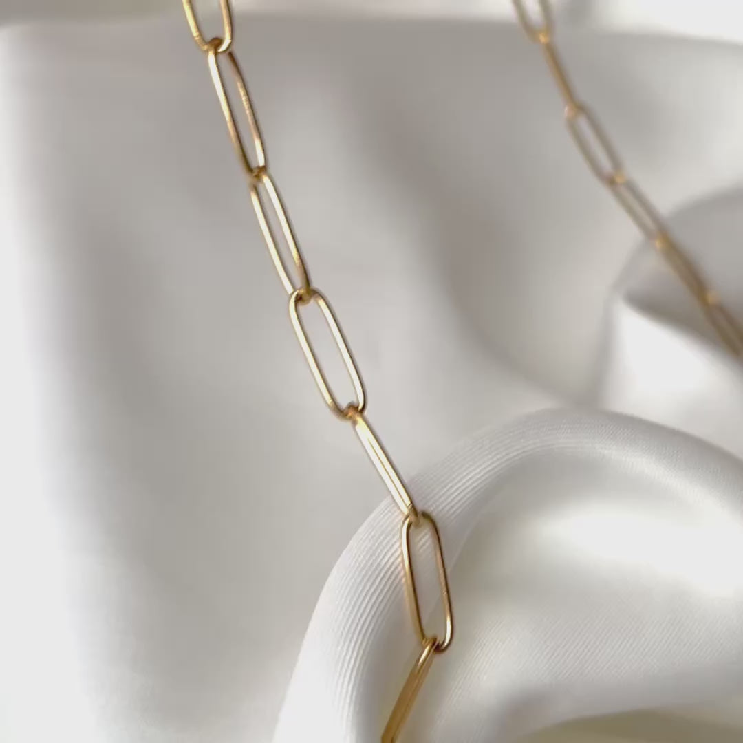 Gold paperclip chain on white satin, links reflecting light as camera changes its angle.