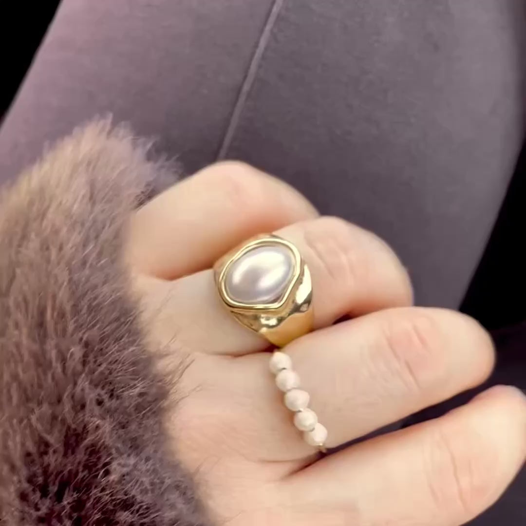 A hand in fist poze wearing dark cool brown fuzzy sleeve and a cool brown pants on the background, with the hand wearing a chunky pearl ring with an organic shaped band in ring finger, and a slim freshwater pearl beaded ring on the mid finger. The hand moves and and the gold bands catch light  as the hand moves.