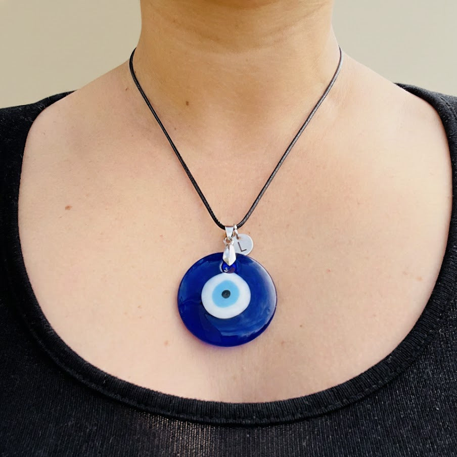 Person wearing an oversized medallion necklace featuring a blue glass evil eye and a tiny initial disc with 'L'