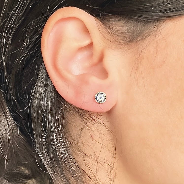 Close-up of an ear wearing tiny round gold stud earrings with blue and clear CZ stones