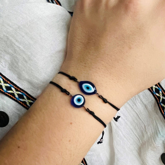 Close-up of a wrist wearing two black evil eye bracelets, one with round one with eye-shaped glass charms on a white fabric background.