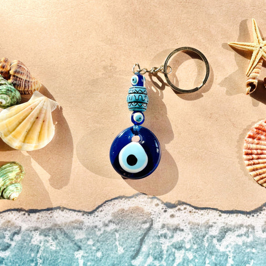 Glass Evil eye keychain with blue bead accents, with seashells and starfish on a sandy beach